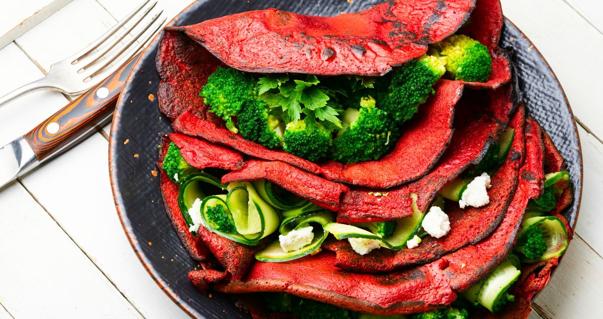 imagem de panquecas de beterraba, que tem a coloração avermelhada, recheadas com brócolis e abobrinha, postas em um prato escuro sobre uma mesa branca de madeira com um garfo e uma faca.