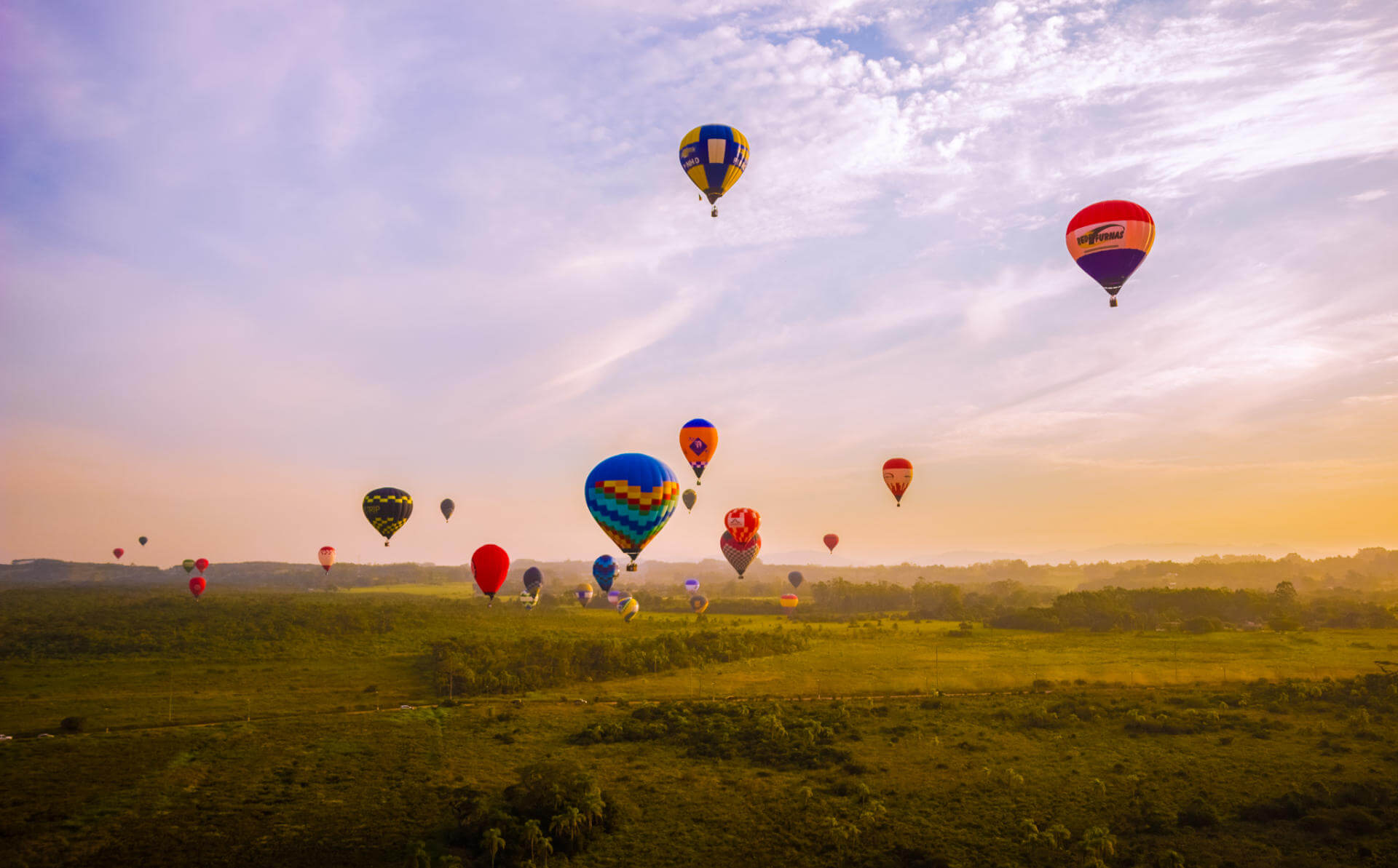 a imagem contempla um horizonte com céu azul alaranjado e vários balões de balonismo no céu, eles são bem coloridos e estão acima de um vasto campo verde.