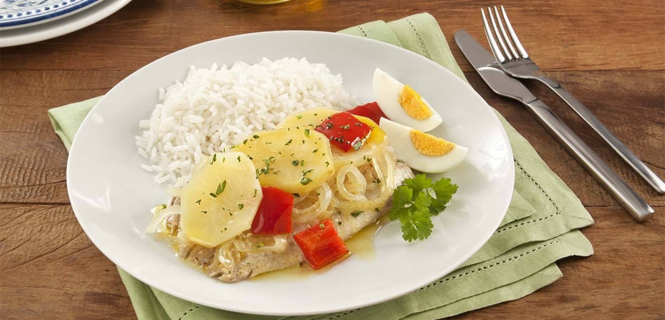 imagem de um prato branco posto sobre uma mesa de madeira. No prato está servido um filé de peixe coberto por pimentões, ovos cozidos e batatas.