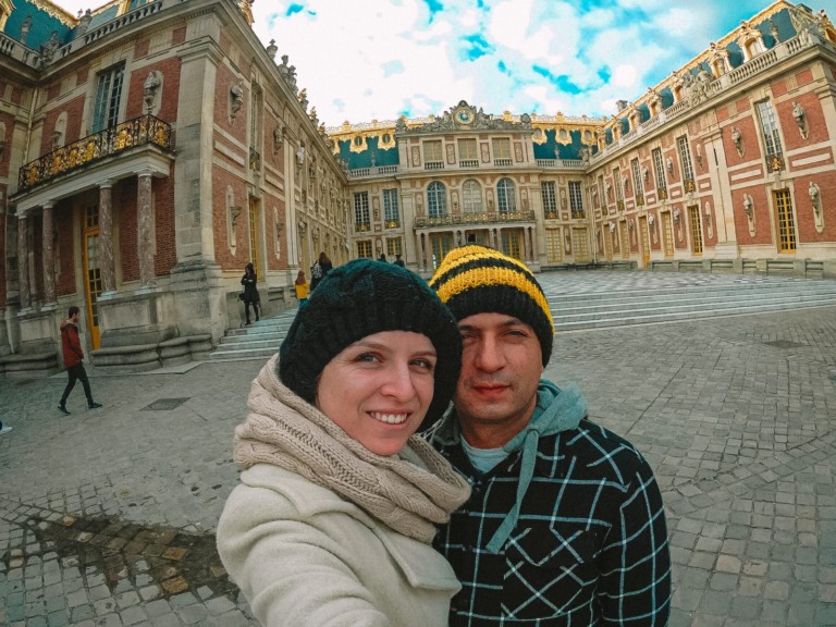 um casal tirando uma selfie em frente ao patio do pal&aacute;cio de versalhes.
