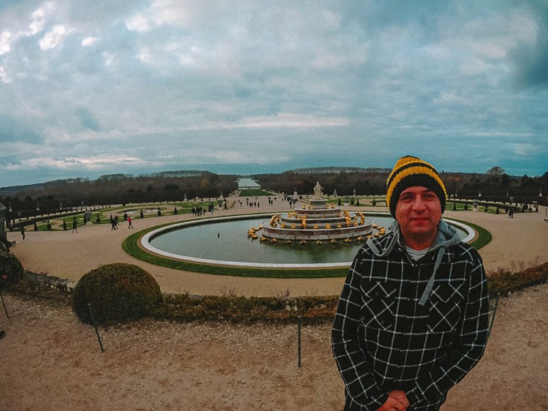 em primeiro plano um homem de casaco preto com xadrez branco e uma touca. Ao fundo um chafariz que marca o inicio do jardim do pal&aacute;cio de versalhes que fica a perder de vista ao final da foto.