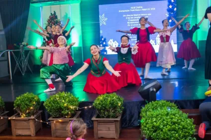 Grupo de dançarinas em trajes natalinos se apresentando em um palco decorado, com árvores de Natal ao fundo.