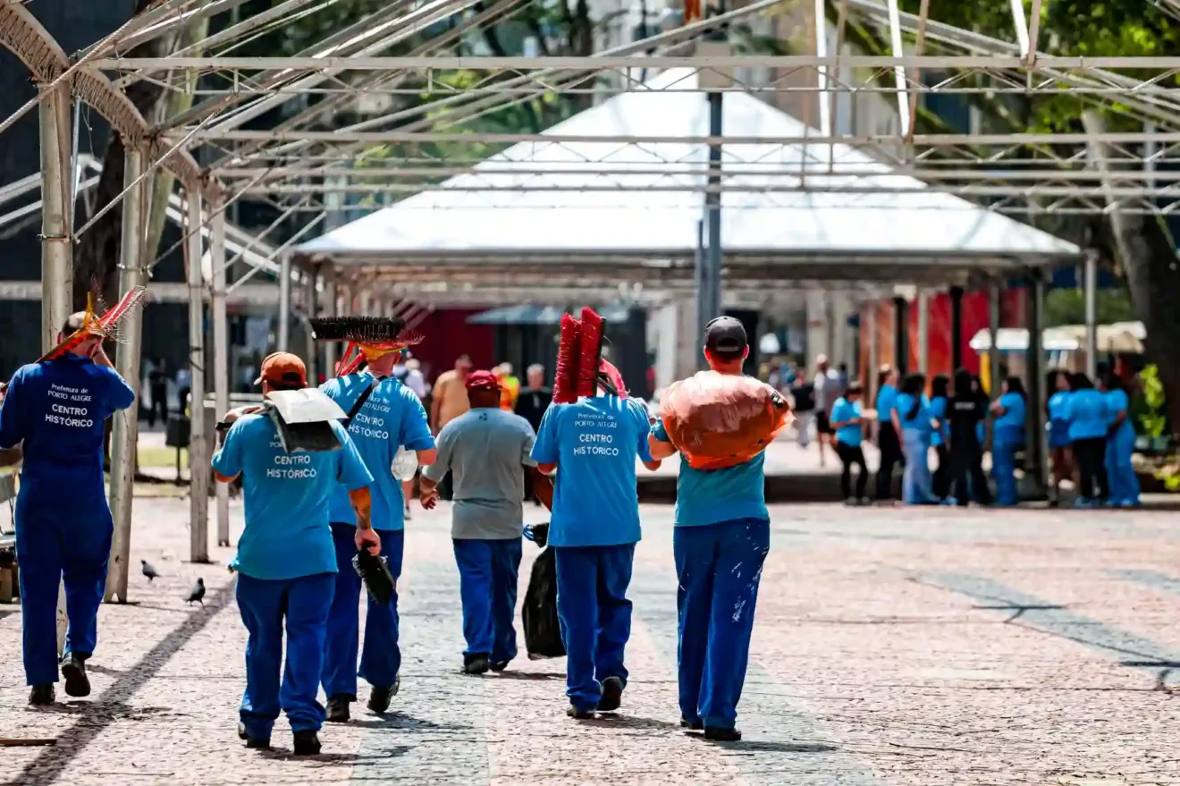 Grupo de trabalhadores em camisetas azuis caminhando pelo Centro Histórico de Porto Alegre, carregando equipamentos e utensílios de varrição.