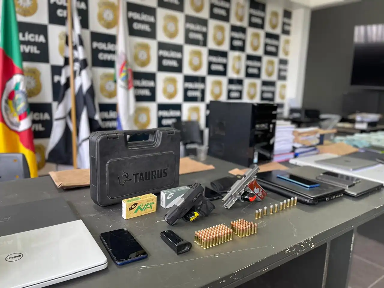 Mesa com material apreendido pela polícia, incluindo armas, munições, e um laptop, em um ambiente de delegacia.