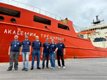 Equipe posando em frente ao navio Akademik Treshnikov, com seis membros em camisetas azuis, no porto.