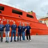 Equipe posando em frente ao navio Akademik Treshnikov, com seis membros em camisetas azuis, no porto.