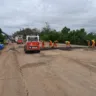 Trabalhadores em obras no quilômetro 46 da FreeWay, com máquinas e equipamentos de construção em um dia nublado.