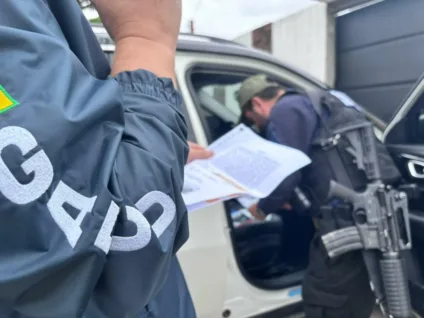 Agentes da polícia em operação, com documentos em mãos e armamento visível, abordando um veículo em um ambiente urbano.