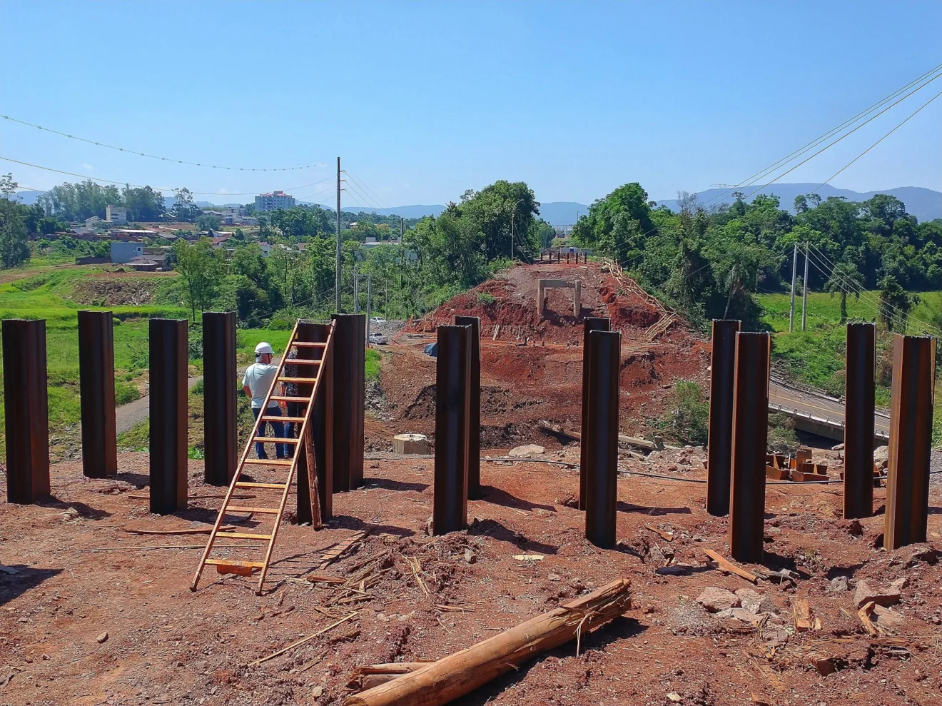 Imagem de uma construção com pilares de aço e uma escada, com uma paisagem verde ao fundo e céu azul.