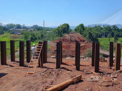 Imagem de uma construção com pilares de aço e uma escada, com uma paisagem verde ao fundo e céu azul.