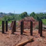 Imagem de uma construção com pilares de aço e uma escada, com uma paisagem verde ao fundo e céu azul.