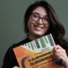 A escritora Tatiane Takahashi Nunes segura o livro O Submarino Quadrado com seu Periscópio Listrado. Ela está com uma blusa preta e óculos, sorrindo para a foto. Ao fundo, é possível ver uma parede verde escuro.