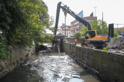 Máquina retroescavadeira realizando limpeza no arroio Passo das Pedras, cercada por vegetação e edifícios ao fundo.