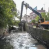 Máquina retroescavadeira realizando limpeza no arroio Passo das Pedras, cercada por vegetação e edifícios ao fundo.