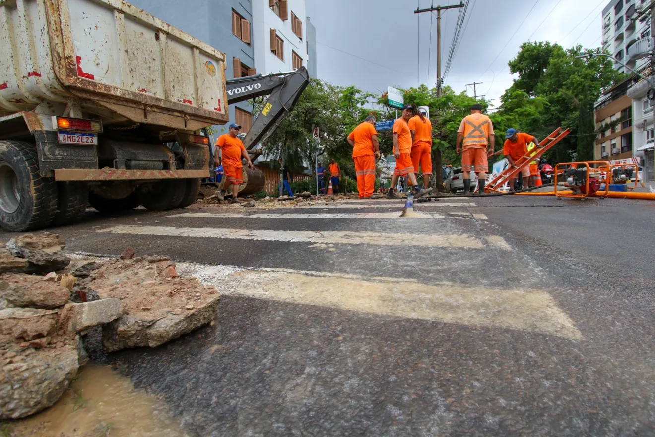 Trabalhadores em uniforme laranja realizando reparo de adutora na avenida Coronel Lucas de Oliveira, em Porto Alegre, com caminhão e máquinas.