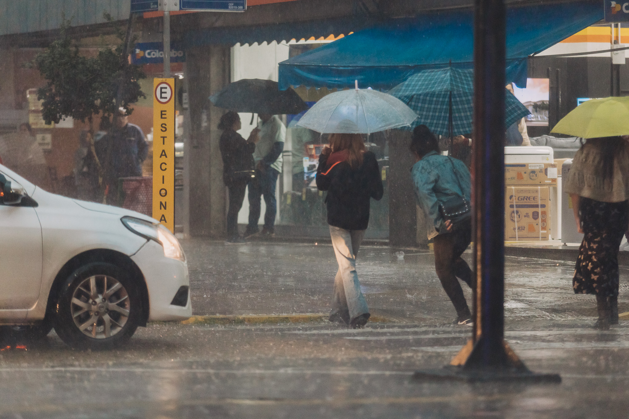 Pessoas caminhando na chuva com guarda-chuvas, em uma rua com trânsito e clima nublado. Imagem demonstra um dia chuvoso na cidade.