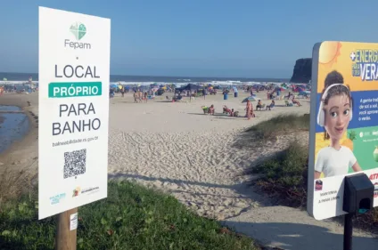 Placa indicando local apropriado para banho na praia, com pessoas e guarda-sóis ao fundo.