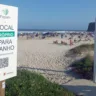 Placa indicando local apropriado para banho na praia, com pessoas e guarda-sóis ao fundo.