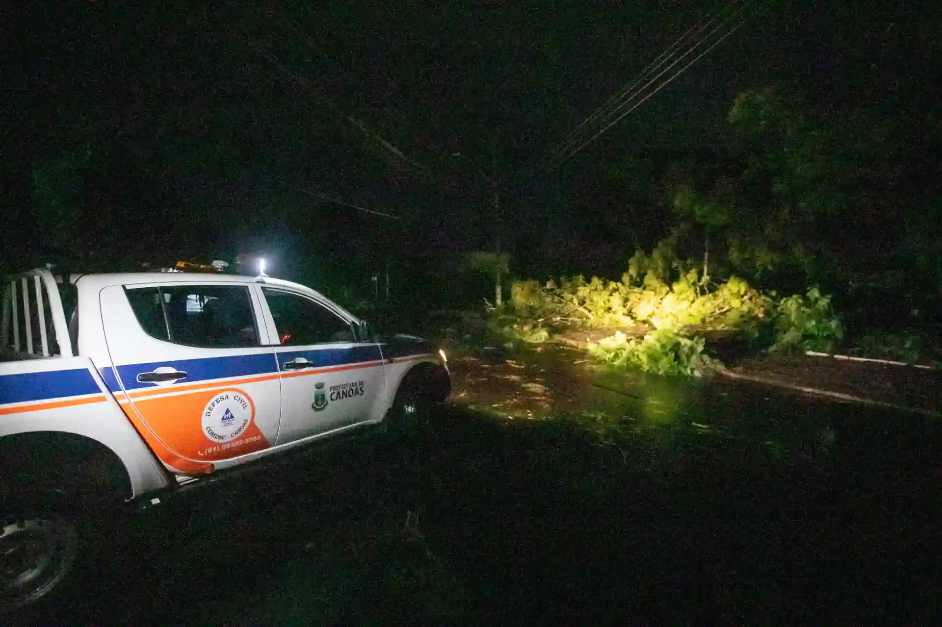 Carro da Defesa Civil de Canoas em uma rua escura, com galhos de árvore caídos e iluminação artificial.