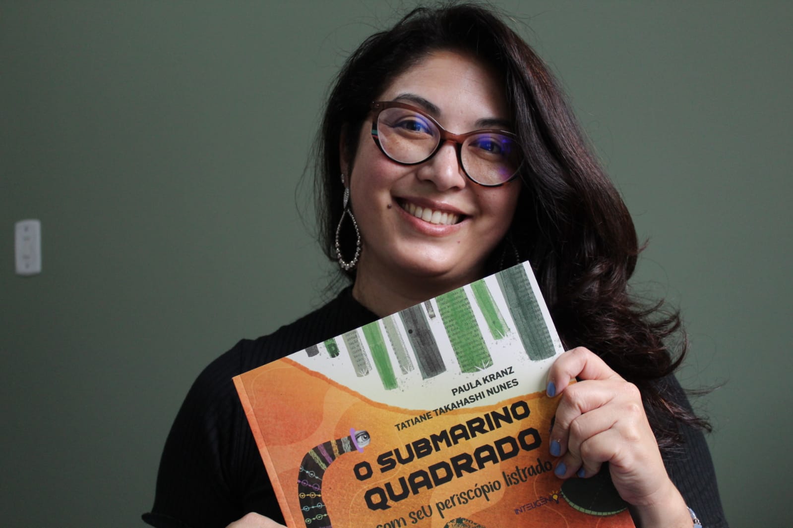 A escritora Tatiane Takahashi Nunes segura o livro O Submarino Quadrado com seu Periscópio Listrado. Ela está com uma blusa preta e óculos, sorrindo para a foto. Ao fundo, é possível ver uma parede verde escuro.