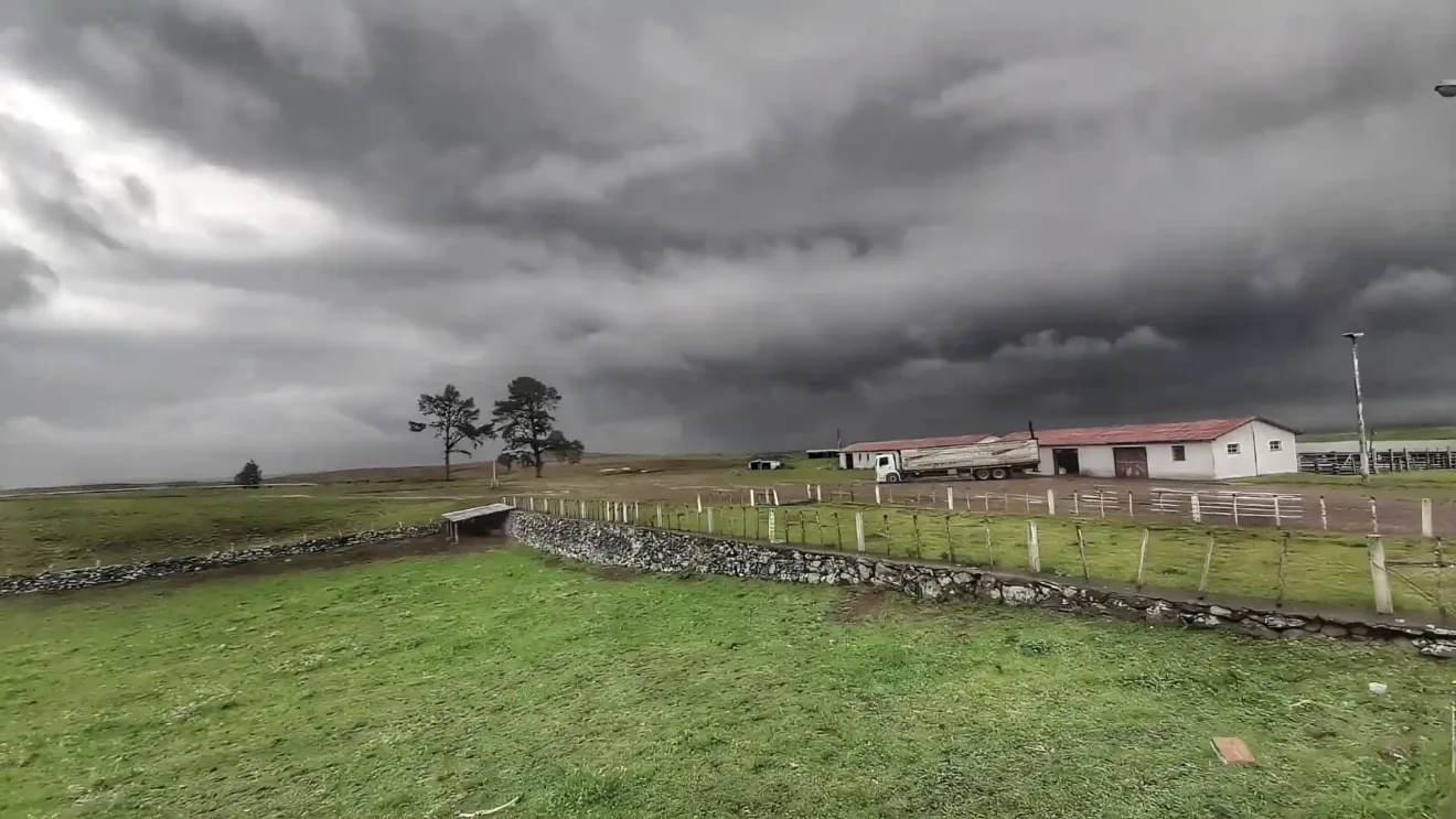 Vista de um campo com céu nublado e escuro, comumente associado a tempestades, cercas de madeira, um galpão e árvores ao fundo.