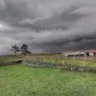 Vista de um campo com céu nublado e escuro, comumente associado a tempestades, cercas de madeira, um galpão e árvores ao fundo.