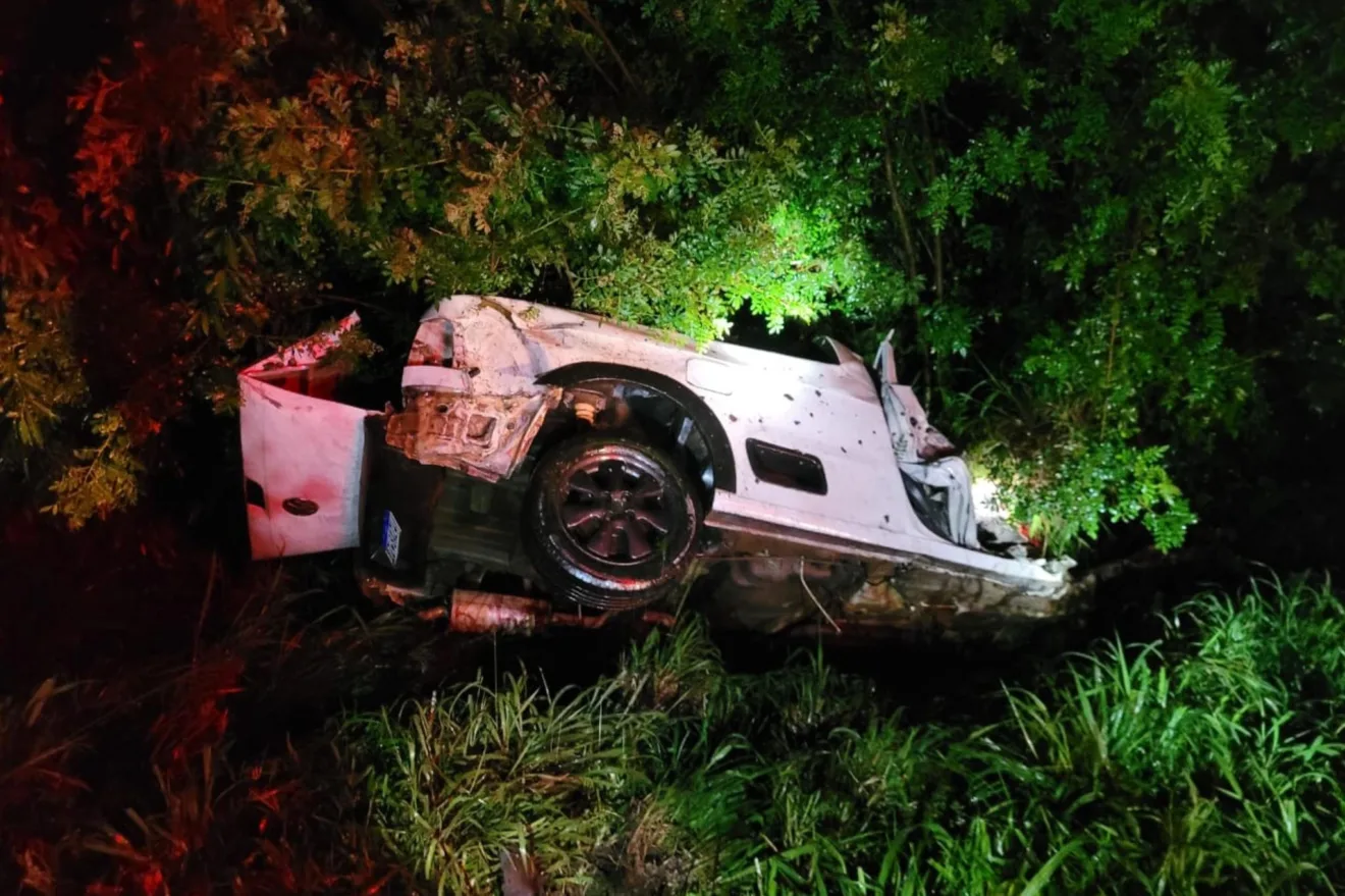 Carro acidentado na BR-153 em Cachoeira do Sul, virado de lado entre vegetação à noite, com danos visíveis.