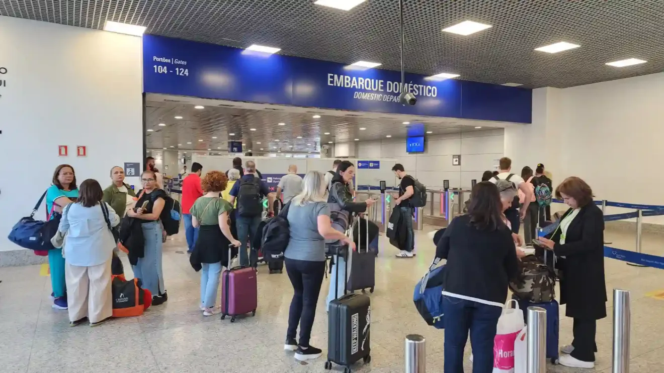 Passageiros aguardando embarque doméstico no Aeroporto Salgado Filho. Eles transportam malas e mochilas e o ambiente está movimentado.