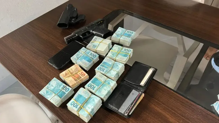 Imagem de uma mesa com pacotes de dinheiro, uma arma e carteiras. Materiais foram apreendidos na Opera&ccedil;&atilde;o Carta Branca.