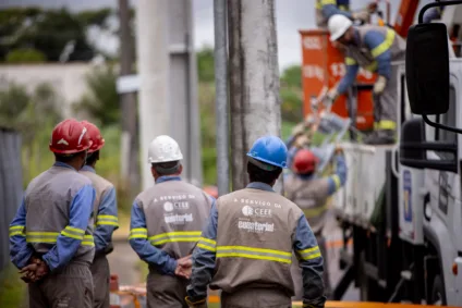 Técnicos da CEEE Equatorial realizando serviços de manutenção em poste. Profissionais vestem capacetes e uniformes.