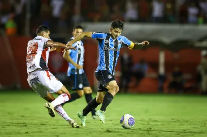 Jogador do Grêmio em ação em uma partida de futebol, destacando-se com a bola enquanto um adversário tenta defender.