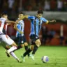 Jogador do Grêmio em ação em uma partida de futebol, destacando-se com a bola enquanto um adversário tenta defender.