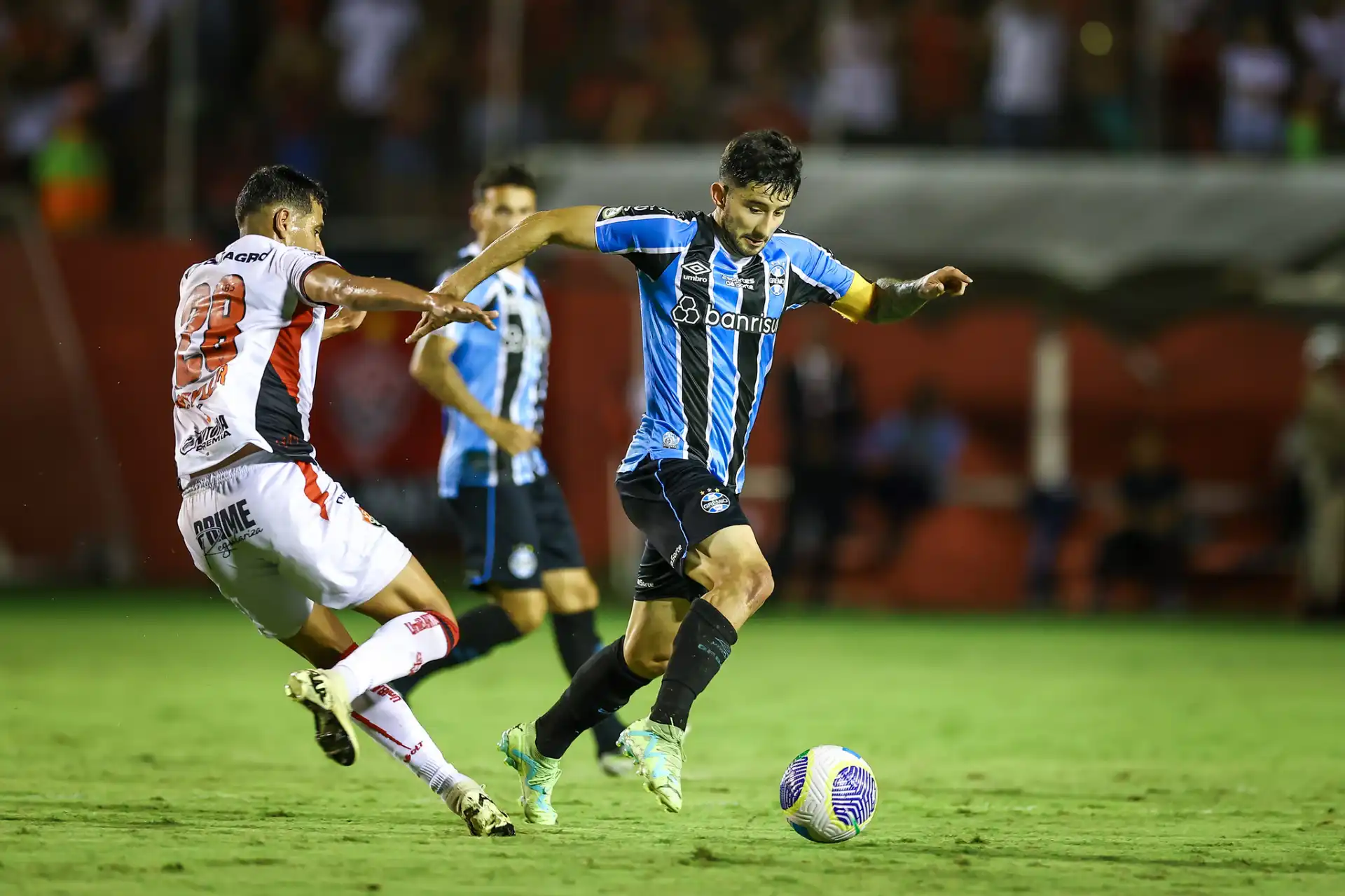 Jogador do Grêmio em ação em uma partida de futebol, destacando-se com a bola enquanto um adversário tenta defender.