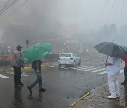 Cena de desastre em Gramado após queda de avião. Pessoas se protegem da chuva enquanto observam os danos e fumaça no local.