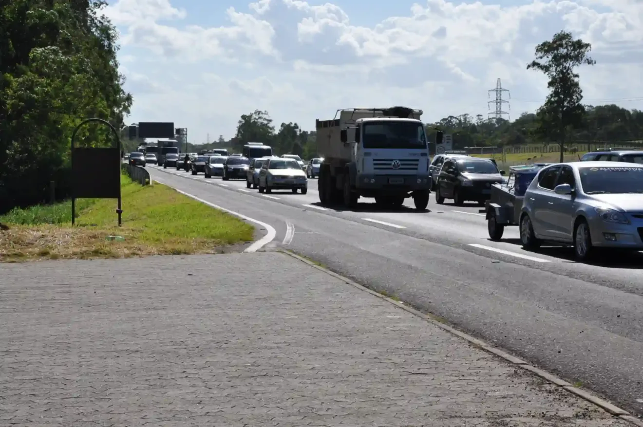 Imagem de uma rodovia movimentada, com vários carros e um caminhão, em um dia ensolarado. A cena mostra a intensa circulação de veículos em direção oposta na pista.