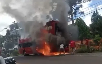 Imagem de um ônibus em chamas, com fumaça e pessoas ao redor tentando apagar o fogo. A cena transmite uma emergência e um momento de tensão.