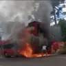 Imagem de um ônibus em chamas, com fumaça e pessoas ao redor tentando apagar o fogo. A cena transmite uma emergência e um momento de tensão.