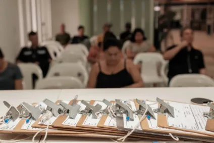 Imagens de pastas e pranchetas dispostas em uma mesa, enquanto pessoas aguardam em cadeiras ao fundo.