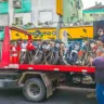 Caminhão em uma rua urbana transportando várias motos apreendidas, com pessoas fazendo o carregamento e um comércio ao fundo.