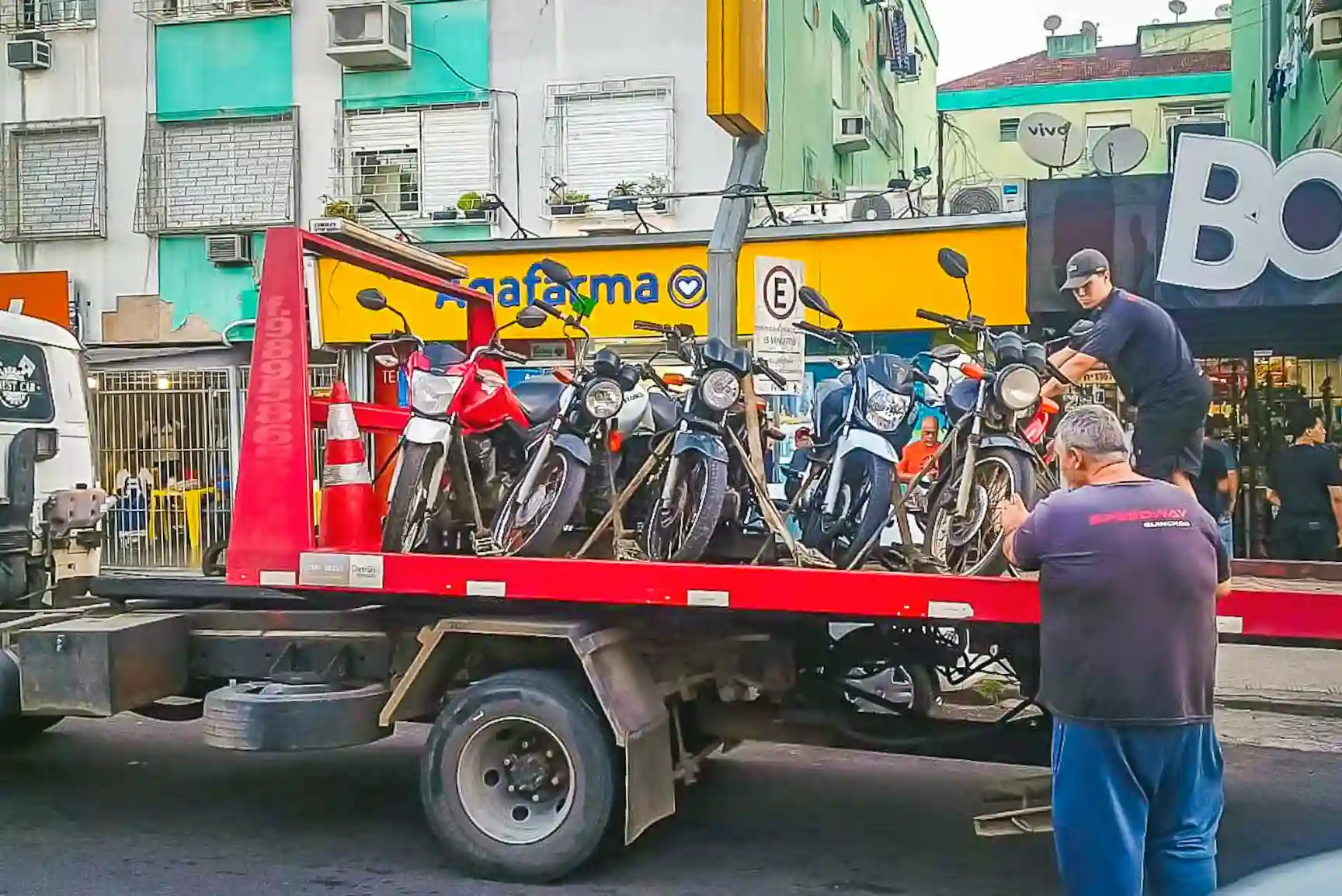 Caminhão em uma rua urbana transportando várias motos apreendidas, com pessoas fazendo o carregamento e um comércio ao fundo.