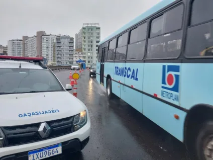 Imagem de um ônibus da Transcal e um veículo da fiscalização em uma rua, cercados por cones de sinalização. O céu está nublado e há prédios ao fundo.