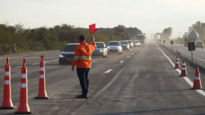 Imagem de um agente de trânsito orientando carros em uma estrada com cones de sinalização. O agente segura uma bandeira vermelha em meio ao tráfego.