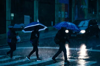 Pessoas atravessando a rua em um dia chuvoso, com guarda-chuvas em meio à chuva forte e reflexos na calçada molhada.