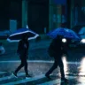 Pessoas atravessando a rua em um dia chuvoso, com guarda-chuvas em meio à chuva forte e reflexos na calçada molhada.