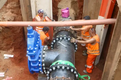 Trabalhadores em um buraco de obra instalando seção de uma adutora, usando equipamentos de segurança e ferramentas.