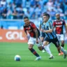 Jogadores do Grêmio e São Paulo disputando a bola em um jogo de futebol, com torcida ao fundo.