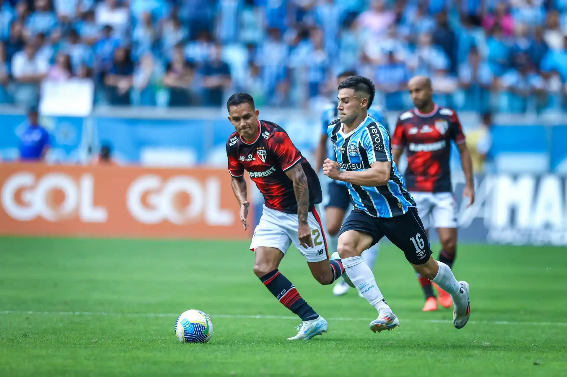 Jogadores do Grêmio e São Paulo disputando a bola em um jogo de futebol, com torcida ao fundo.