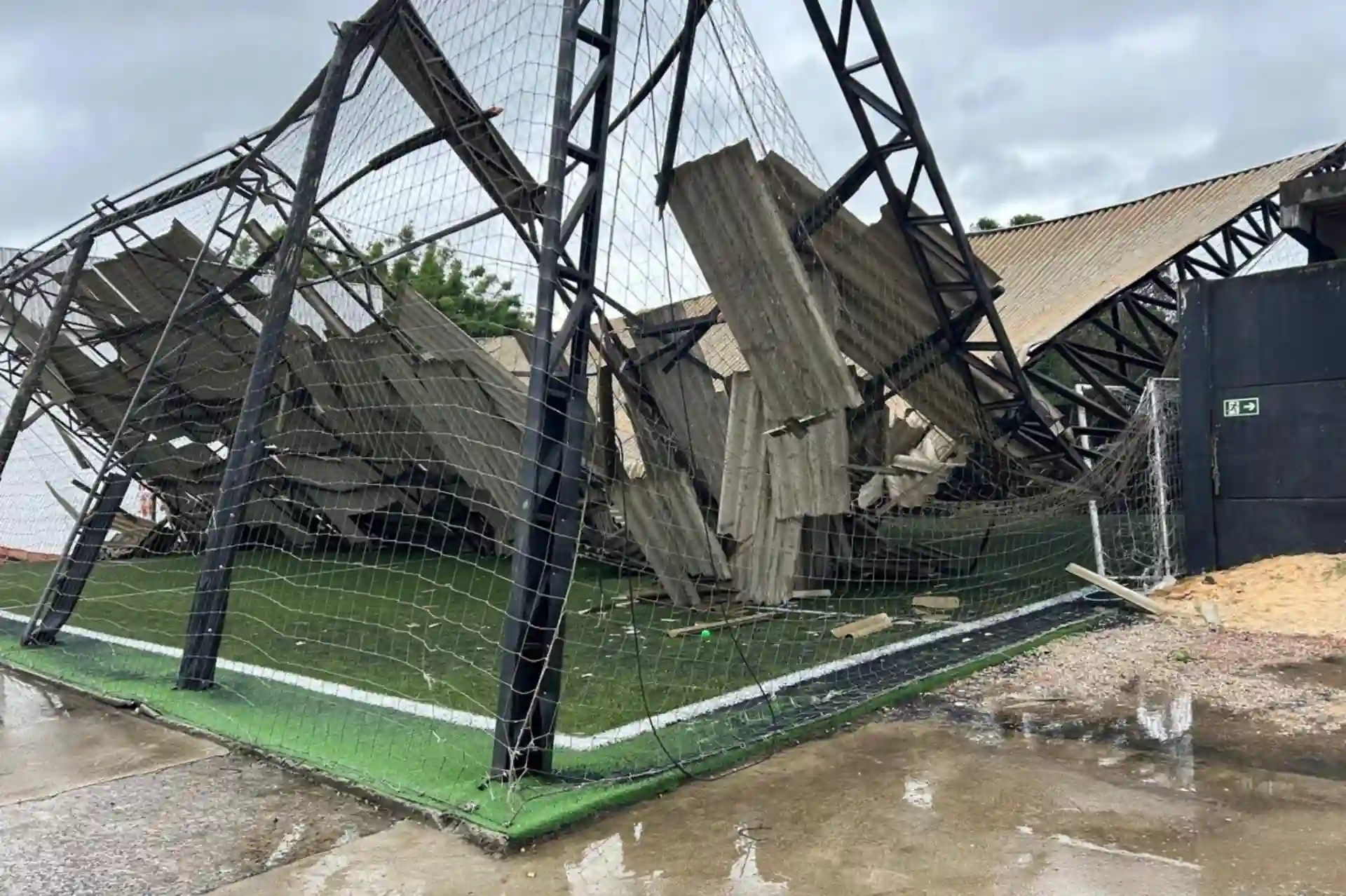 Imagem de um campo de futebol com a estrutura de arquibancada danificada. A cena mostra os destroços e a rede, evidenciando os danos causados pela intempérie.