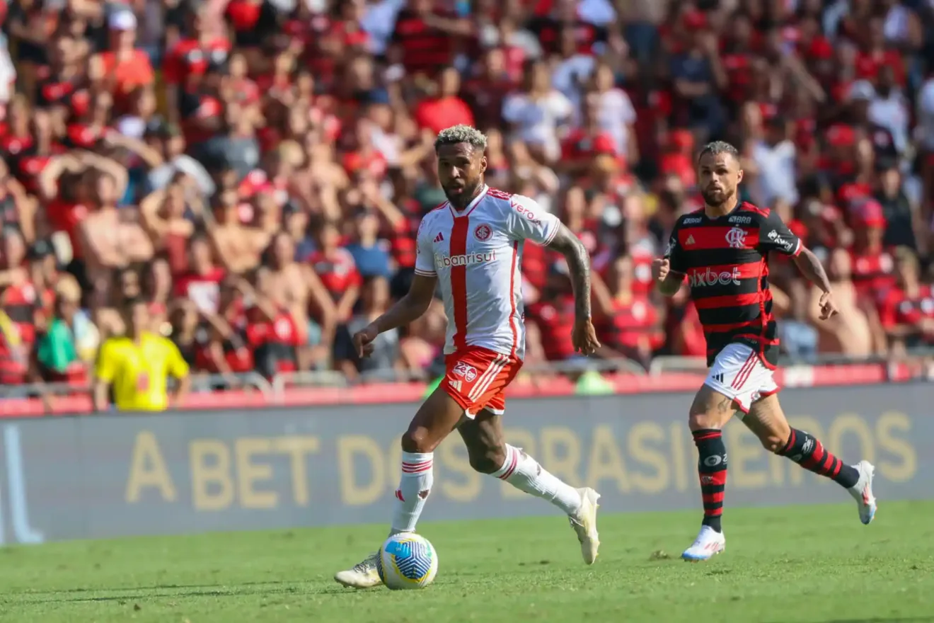 Jogadores do Internacional e Flamengo em campo durante uma partida de futebol, com torcedores ao fundo.