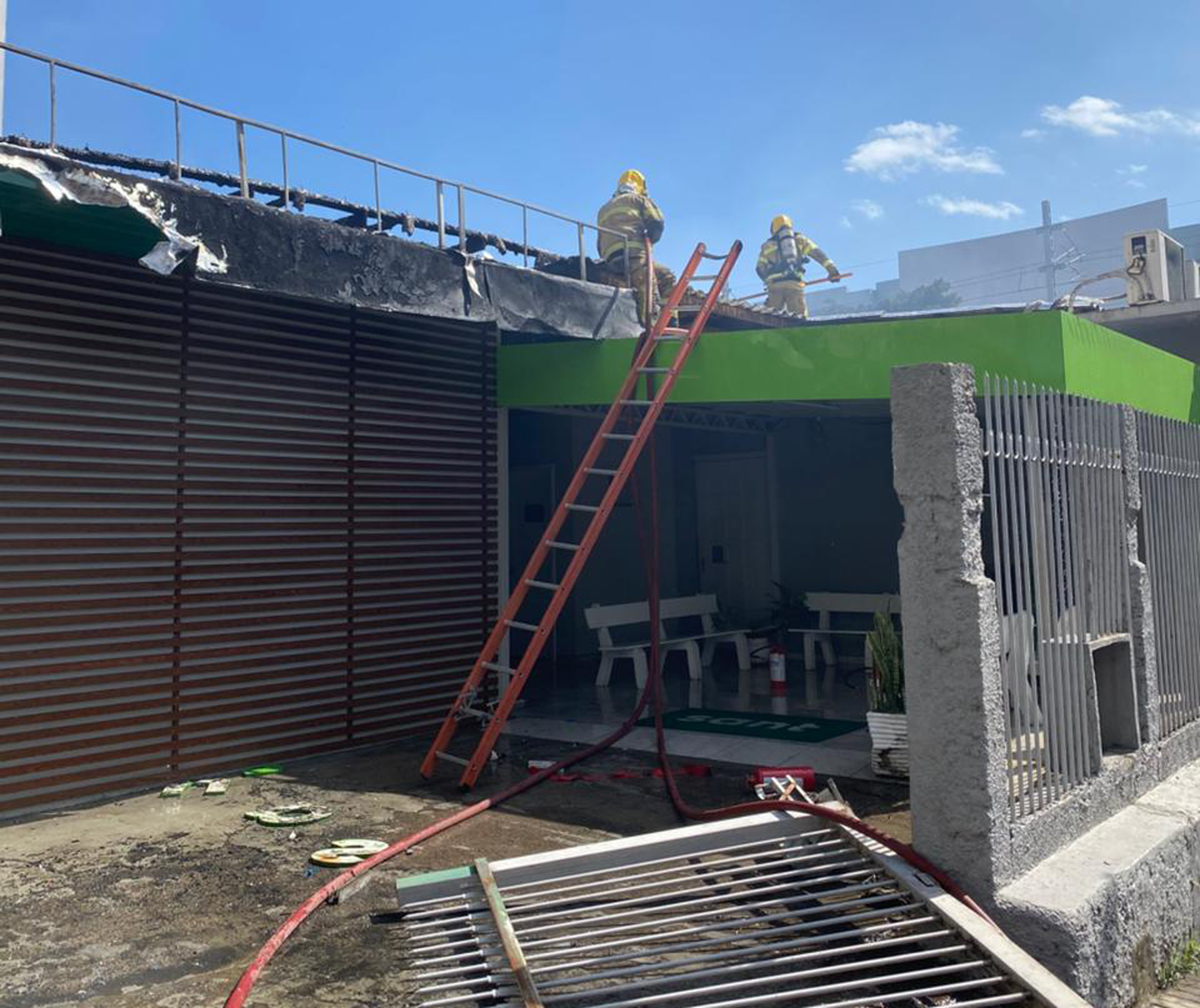 Dois bombeiros atuando no combate a incêndio em um telhado. Eles utilizam equipamentos de segurança e estão em uma escada. Céu azul ao fundo.